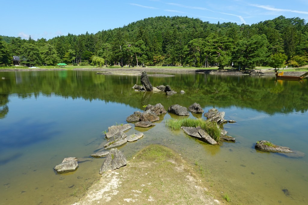 The large pond of Motsu-ji's Pure Land garden in Hiraizumi, with a low peninsula and scattered rocks in the water and wooded hills reflected behind