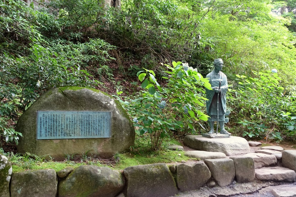 Bronze statue of the haiku poet Matsuo Basho in traveling clothes, standing among green trees beside a large stone with a plaque on the grounds of Chuson-ji