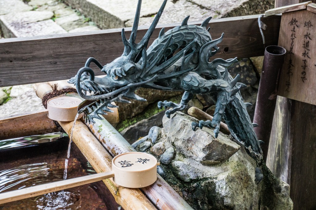 A bronze dragon spout pouring water into a stone purification basin, with wooden ladles laid across bamboo