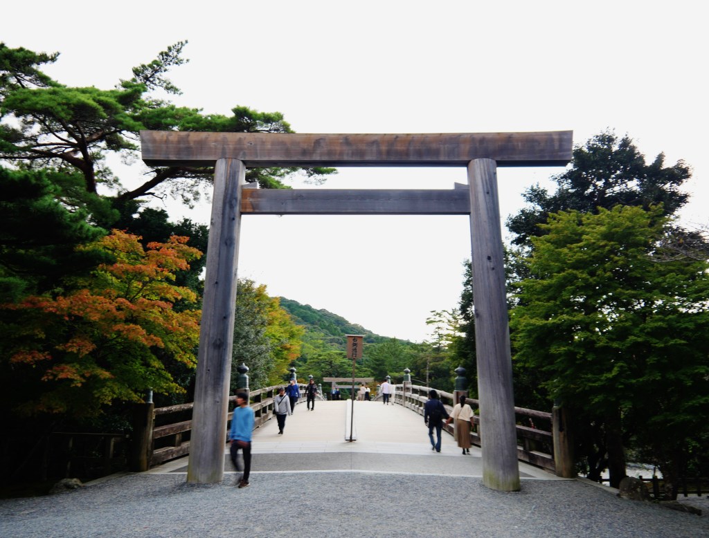 A plain unpainted wooden torii with a straight top beam at the entrance to the Uji Bridge of Ise Jingu, with forested hills beyond