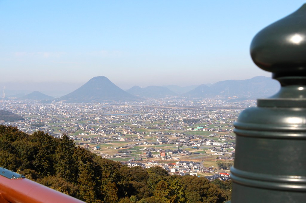 View from the mountainside at Kotohira-gu over the flat, built-up Sanuki plain, with a cone-shaped hill, Mount Iino, rising on the horizon