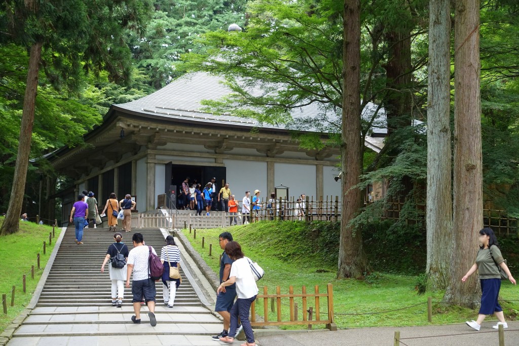 Visitors climbing stone steps toward the modern outer hall that houses the Konjikido at Chuson-ji, among tall cedar trees