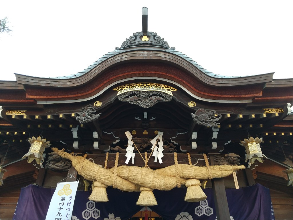 The front of a shrine worship hall with a thick twisted straw shimenawa rope and white zigzag paper shide hanging beneath an ornate curved roof